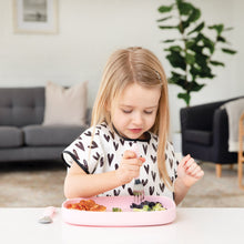 Load image into Gallery viewer, A blonde girl in a black &amp; white bib eats a meal from a Bumkins Silicone Grip Tray: Pink with pink utensils at a white table.
