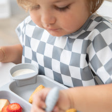 Load image into Gallery viewer, A toddler wearing a Bumkins Junior Bib: Charcoal Check focuses on eating from a divided tray while sitting at a white table.
