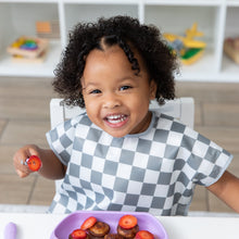 Load image into Gallery viewer, A child smiling, wearing Bumkins Junior Bib: Charcoal Check at a white table while eating pancakes and strawberries from a purple plate.
