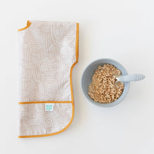 Load image into Gallery viewer, Bumkins Junior Bib: Wander with a catch-all pocket lays folded next to a bowl of cereal and baby spoon on a white background.

