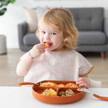 Load image into Gallery viewer, A blonde toddler eats from an orange plate at a white table, wearing a Bumkins Junior Bib: Wander. A gray sofa is in the background.
