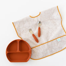 Load image into Gallery viewer, Bumkins Junior Bib: Wander lays flat on a white background with orange utensils on top of it, next to an orange divided plate.
