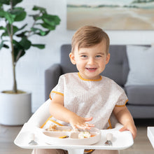 Load image into Gallery viewer, A toddler beams in a high chair while wearing a Bumkins Junior: Bib Wander, eating food from a beige divided plate.
