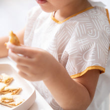 Load image into Gallery viewer, Close-up of a child wearing a Bumkins Junior Bib: Wander, holding a snack while more pieces fill a beige bowl on the white table.
