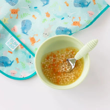 Load image into Gallery viewer, A green bowl of soup with a green spoon sit next to Bumkins Junior Bib: Ocean Life on a white background.
