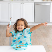 Load image into Gallery viewer, A curly-haired child in a Bumkins Junior Bib: Ocean Life sits at a table with a blue plate, holding a spoon and raising an arm excitedly.
