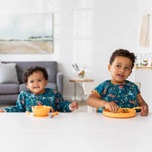 Load image into Gallery viewer, Two happy toddlers in matching outfits sit at a white table eating from orange dishes; one wears a Bumkins Junior Bib: Jungle.
