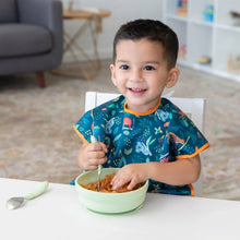 Load image into Gallery viewer, A child in a Bumkins Junior Bib: Jungle smiles, enjoying pasta from a green bowl at a white table.
