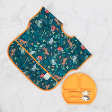 Load image into Gallery viewer, Bumkins Junior Bib: Jungle lays next to orange plate with orange utensils on a marble surface.
