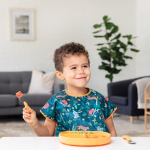 Load image into Gallery viewer, A boy grins, wearing an animal patterned bib as he eats from a Bumkins Silicone Grip Dish: Tangerine with matching utensils.
