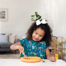 Load image into Gallery viewer, A young girl with curly hair and a bow wearing a Bumkins Junior Bib: Jungle eats from an orange plate using a fork at a white table.
