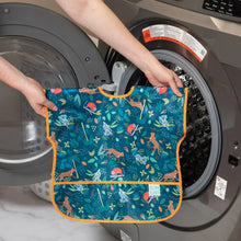 Load image into Gallery viewer, A person holds Bumkins Junior Bib: Jungle in front of an open front-loading washing machine.

