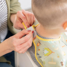 Load image into Gallery viewer, A person fastens the hook-and-loop closure of a Bumkins Junior Bib: Camp Gear around a child's neck.
