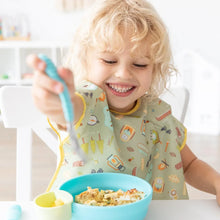 Load image into Gallery viewer, A curly blonde child in a Bumkins Junior Bib: Camp Gear smiles, enjoying a meal from a blue bowl with a fork at a white table.
