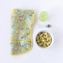 Load image into Gallery viewer, Bumkins Junior Bib: Camp Gear lays folded on a white background next to a green cup, a gray bowl of macaroni and peas, and a fork.
