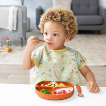Load image into Gallery viewer, A curly-haired child eats from a divided plate with a fork, wearing a Bumkins Junior Bib: Camp Gear at a white table in a cozy living room.
