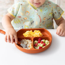 Load image into Gallery viewer, A child wearing a Bumkins Junior Bib: Camp Gear at a white table eats scrambled eggs and fruit from a divided plate using a fork.
