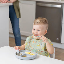 Load image into Gallery viewer, A smiling toddler wears a Bumkins Junior Bib: Camp Gear and eats from a divided plate at a table as an adult stands nearby in a kitchen.
