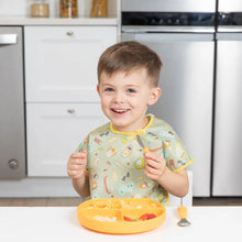 Load image into Gallery viewer, A smiling boy sits at a white table with a yellow plate, wearing a Bumkins Junior Bib: Camp Gear while holding a fork.
