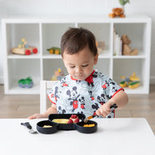 Load image into Gallery viewer, A toddler wearing a Bumkins Junior Bib: Mickey Mouse Classic eats with a fork at a white table, with toys and books in the background.
