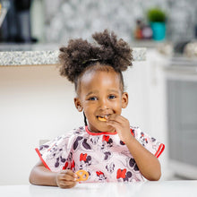 Load image into Gallery viewer, A curly-haired child in Bumkins Junior Bib: Minnie Mouse Classic smiles, eating a pretzel at a white table in a kitchen.
