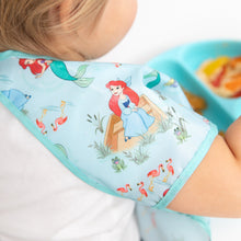 Load image into Gallery viewer, A child dons the Bumkins Junior Bib: Ariel, featuring the red-haired mermaid, next to a blue tray of food.
