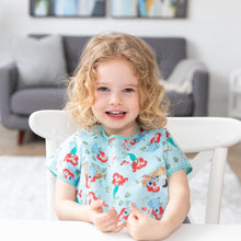 Load image into Gallery viewer, A smiling curly-haired girl at a table, wearing a Bumkins Junior Bib: Ariel with a cozy living room backdrop.
