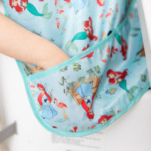 Load image into Gallery viewer, A child places their hand in the pocket of a Bumkins Junior Bib: Ariel featuring colorful cartoon mermaids and flamingos.

