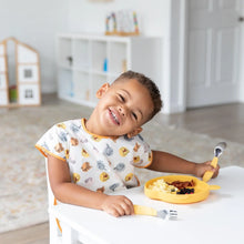 Load image into Gallery viewer, A child wearing a Bumkins Junior Bib: Winnie and Friends eats from a yellow plate at a white table in a bright room.
