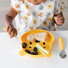 Load image into Gallery viewer, A child eats sandwiches, berries, and celery from a bear-shaped plate while wearing a Bumkins Junior Bib: Winnie and Friends at a table.

