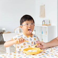 Load image into Gallery viewer, A child with glasses enjoys a meal from a yellow plate with an adult nearby, wearing a Bumkins Junior Bib: Winnie and Friends.
