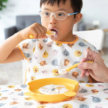 Load image into Gallery viewer, A child in glasses wears a Bumkins Junior Bib: Winnie and Friends while eating yogurt from a yellow bowl with adult assistance.
