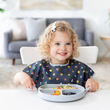 Load image into Gallery viewer, A curly-haired child in a Bumkins Junior Bib: Super Mario™ Lineup sits happily at a white table holding utensils near a gray plate of food.
