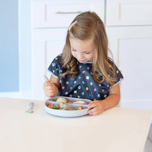 Load image into Gallery viewer, A young girl in a Bumkins Junior Bib: Super Mario™ Lineup sits at a table, eating with utensils from a plate of various foods.

