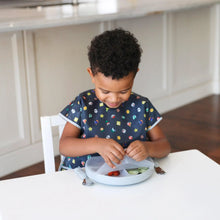 Load image into Gallery viewer, A child smiles while removing a lid from a gray divided plate, wearing a Bumkins Junior Bib: Super Mario™ Lineup with a kitchen visible.
