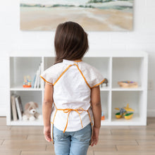 Load image into Gallery viewer, A child in a Bumkins Short-Sleeved Smock: Wander faces a white shelf filled with toys and books, showcasing the neck and waist closures.
