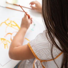 Load image into Gallery viewer, Close-up of a child wearing Bumkins Short-Sleeved Smock: Wander paints in orange and yellow at a white table.
