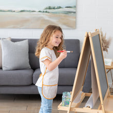 Load image into Gallery viewer, A young girl in a Bumkins Short-Sleeved Smock: Wander paints at an easel, surrounded by a cozy living room with a sofa and landscape art.
