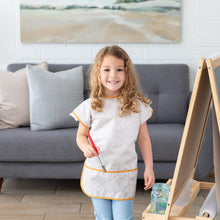 Load image into Gallery viewer, A young girl with curly hair wearing a Bumkins Short-Sleeved Smock: Wander stands in a living room, holding a paintbrush by a wooden easel. 
