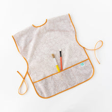 Load image into Gallery viewer, Bumkins Short-Sleeved Smock: Wander lays on a white background with two paintbrushes in the pocket.

