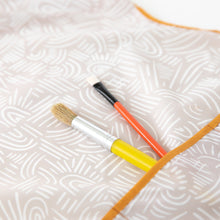 Load image into Gallery viewer, Close-up of two paintbrushes in the pocket of a Bumkins Short-Sleeved Smock: Wander.
