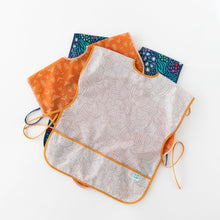 Load image into Gallery viewer, Bumkins Short-Sleeved Smock: Wander lays on top of a stack of similar art smocks on a white background.
