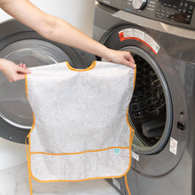 Load image into Gallery viewer, A person holds a Bumkins Short-Sleeved Smock: Wander in front of an open washing machine.
