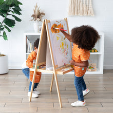Load image into Gallery viewer, Two children, one in a Bumkins Short-Sleeved Smock: Grounded, paint freely on an easel in a bright room with shelves and a plant.
