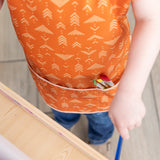 Close-up of a child in a Bumkins Short-Sleeved Smock: Grounded with paintbrushes in the pocket, painting at an easel. 