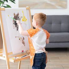 Load image into Gallery viewer, A toddler in a Bumkins Short-Sleeved Smock: Grounded paints at an easel with a paintbrush, with a couch in the background.
