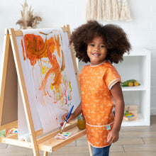Load image into Gallery viewer, A child with curly hair joyfully paints vibrant brushstrokes on an easel, wearing a Bumkins Short-Sleeved Smock: Grounded.
