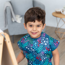 Load image into Gallery viewer, A child beams at the camera in a Bumkins Short-Sleeved Smock: Animal Prints, beside an easel, gray chair, and wooden table.
