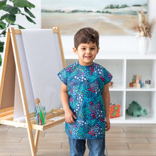 Load image into Gallery viewer, A child in a Bumkins Short-Sleeved Smock: Animal Prints grins next to an easel amid shelves of toys in a bright room.
