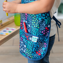 Load image into Gallery viewer, Close-up of a child in a Bumkins Short-Sleeved Smock: Animal Prints next to an easel with a red paintbrush in the pocket.
